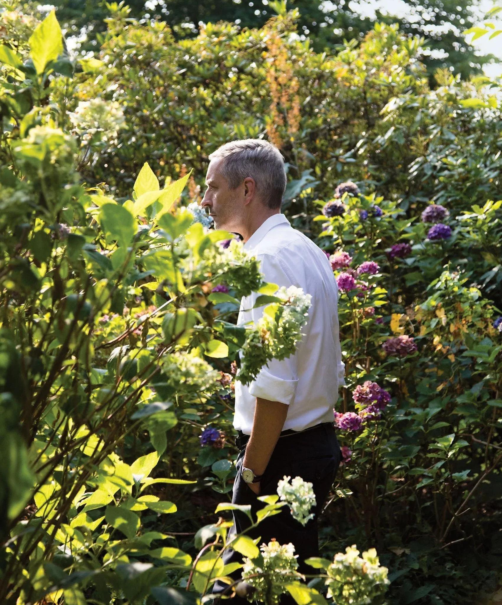Dries Van Noten standing among lush green plants and flowers in a garden, wearing a light shirt and dark trousers.