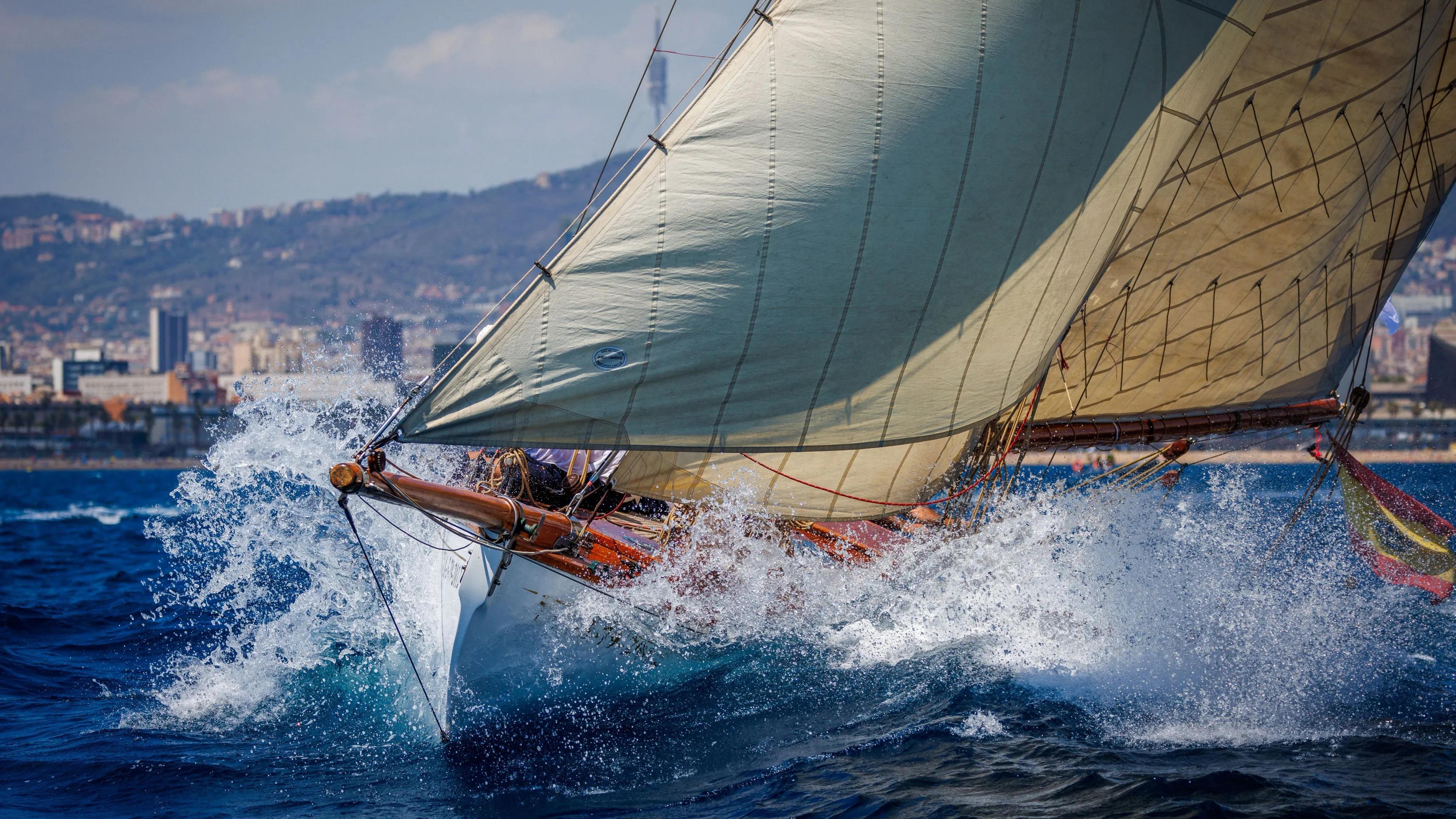 Close-up of a classic sailing yacht cutting through the sea during the Puig Vela Clàssica Barcelona regatta, with waves splashing against its hull and sails.