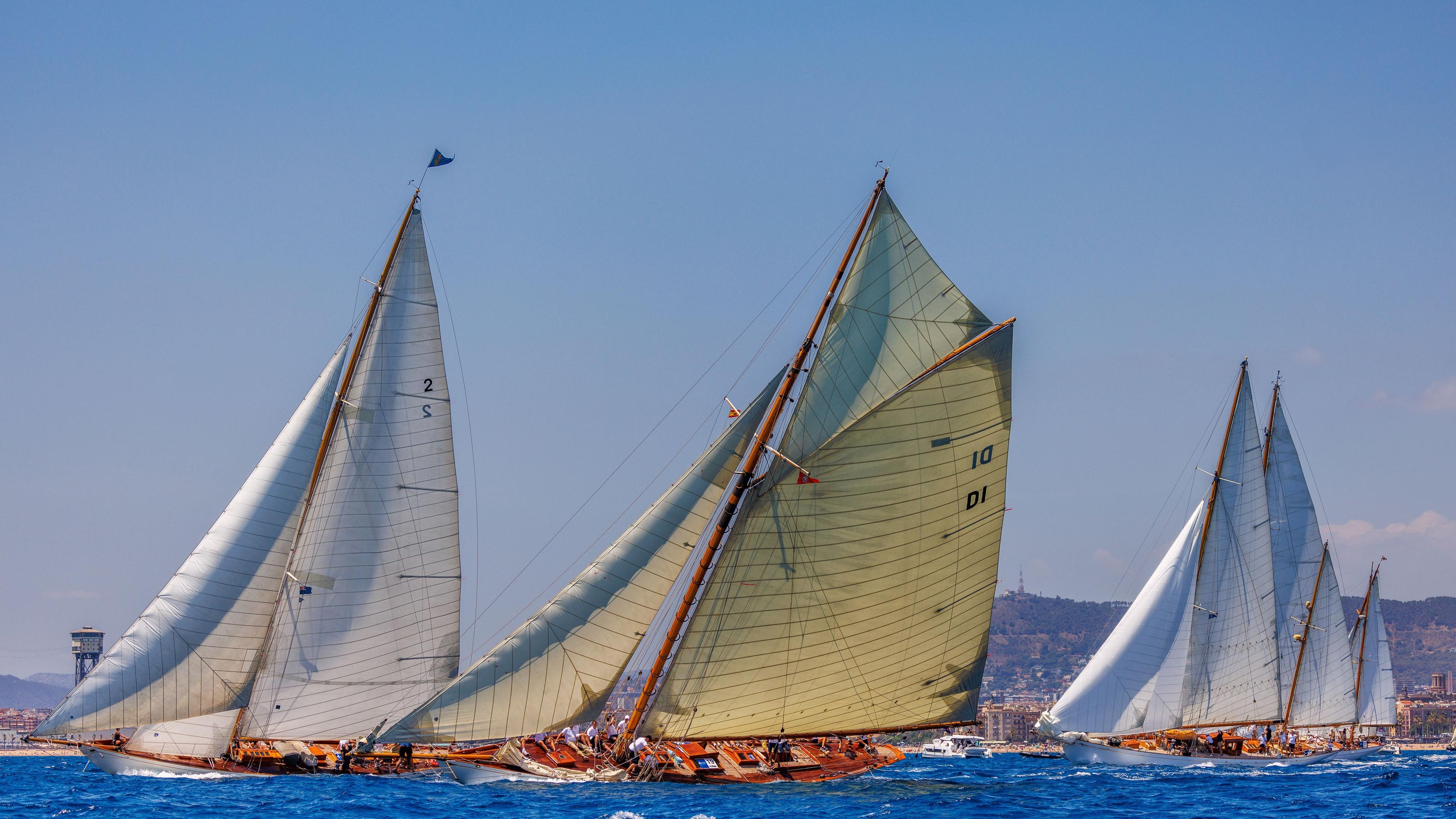 Classic sailboats racing at sea during the Puig Vela Clàssica Barcelona regatta, led by yachts with white and pale green sails.