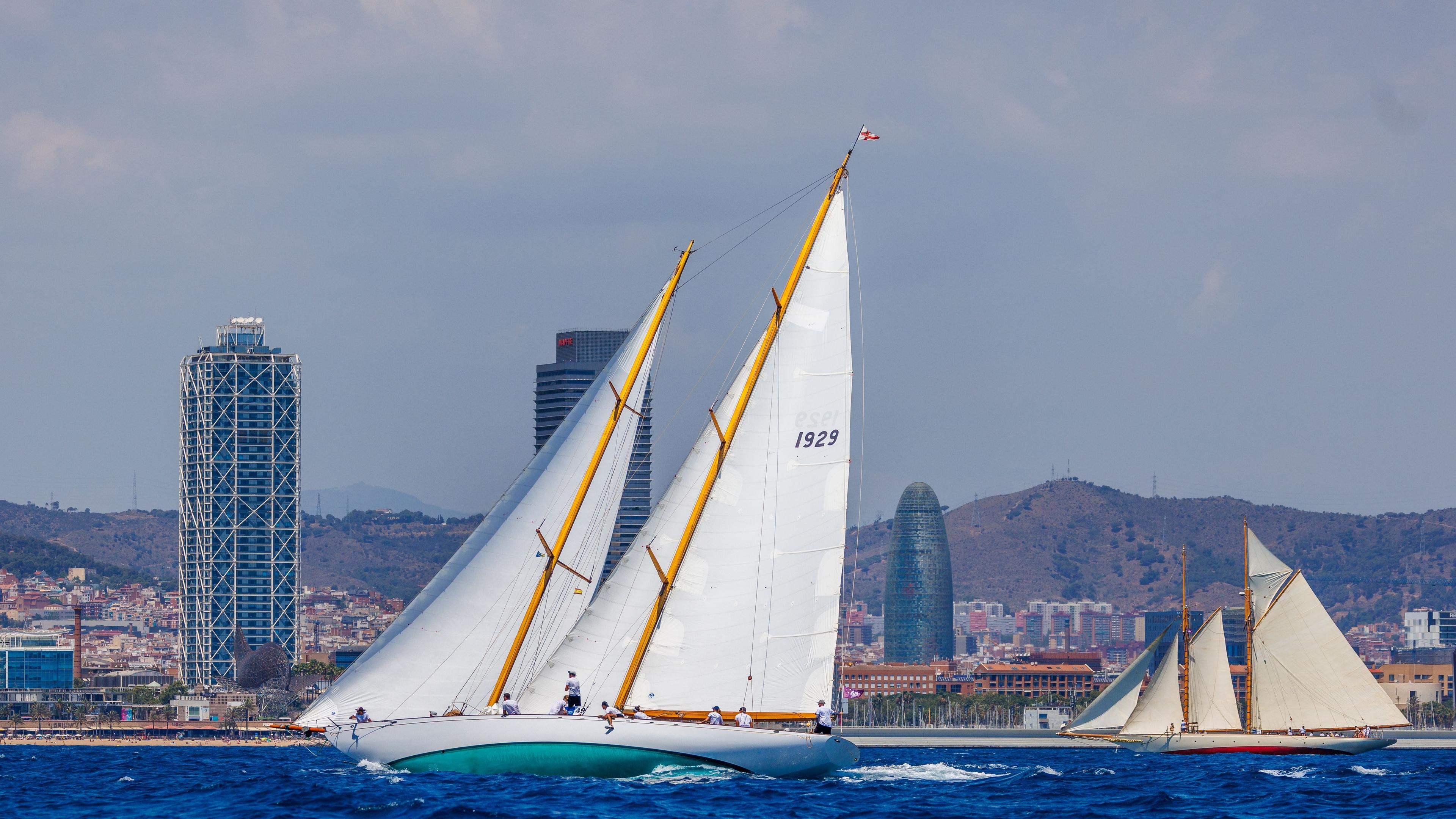 Classic sailing boats racing off the Barcelona coast during the Puig Vela Clàssica Barcelona regatta, with the city skyline in the background.