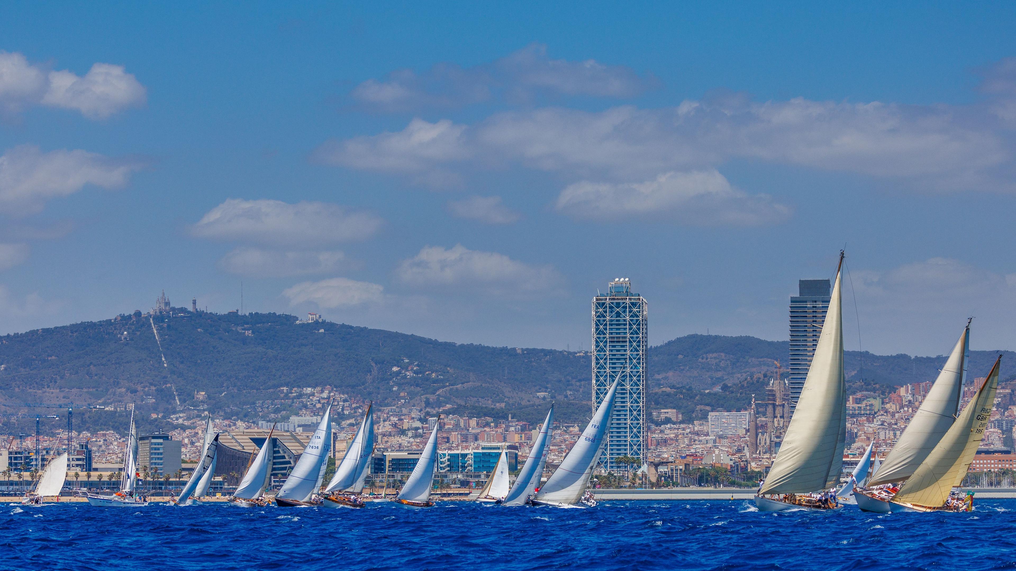 Classic sailboats racing off the Barcelona coast during the Puig Vela Clàssica Barcelona regatta, with the city skyline and mountains in the background.