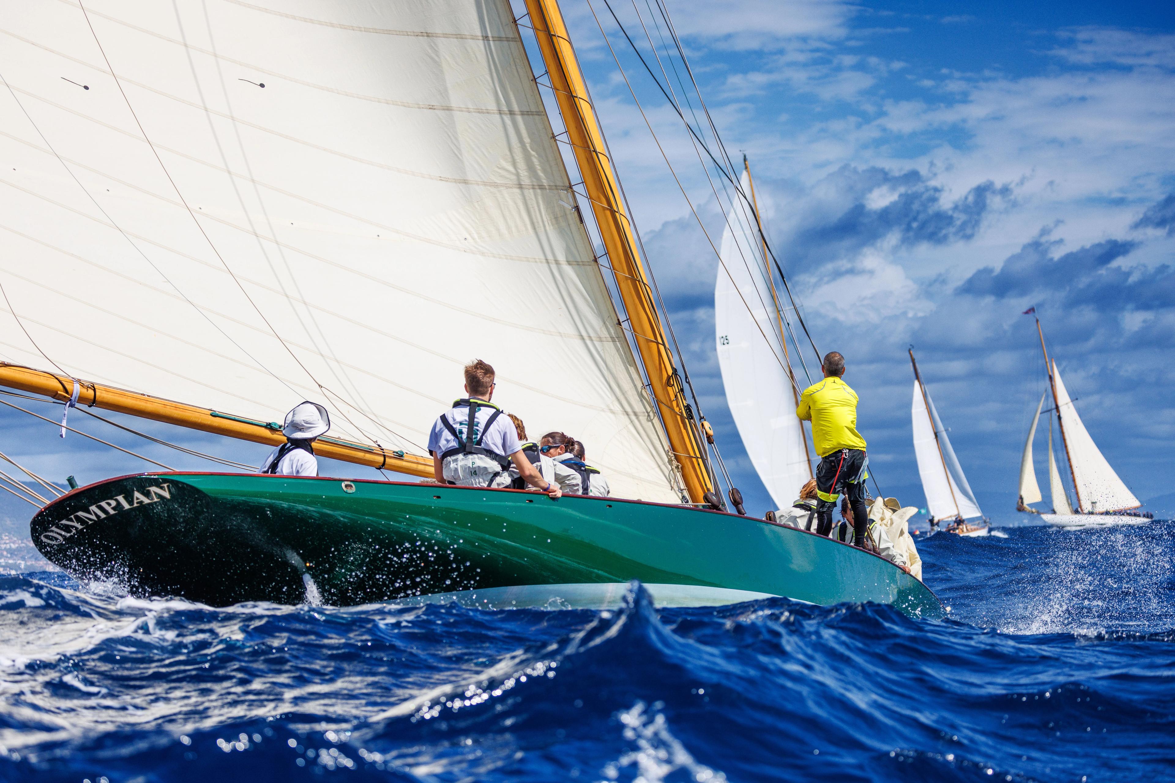 Classic sailboats racing at sea during the Puig Vela Clàssica Barcelona regatta, with crew members aboard a green-hulled yacht in the foreground.