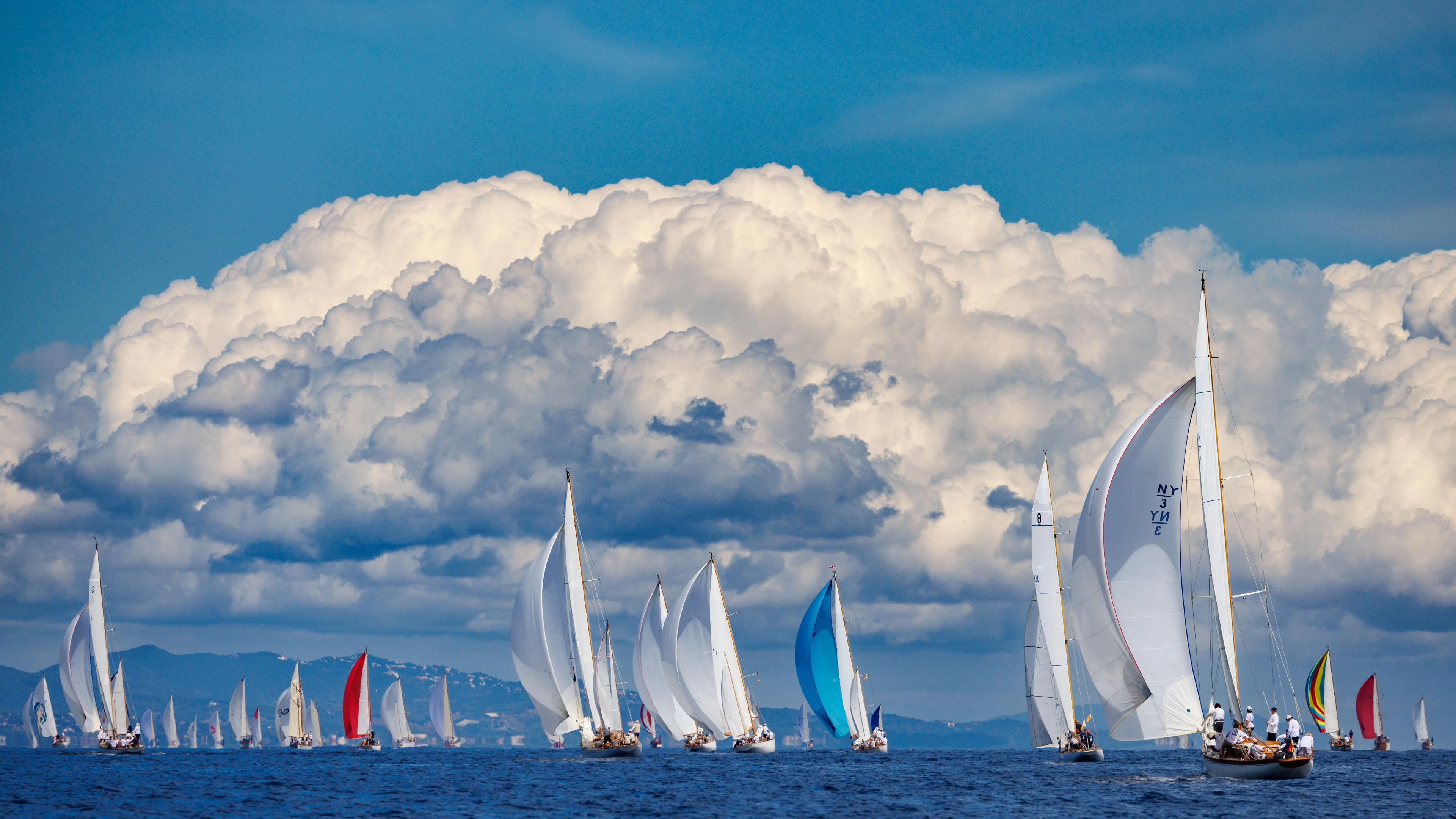 Sailboats racing on the sea during the Puig Vela Clàssica Barcelona regatta, with large clouds in the background.