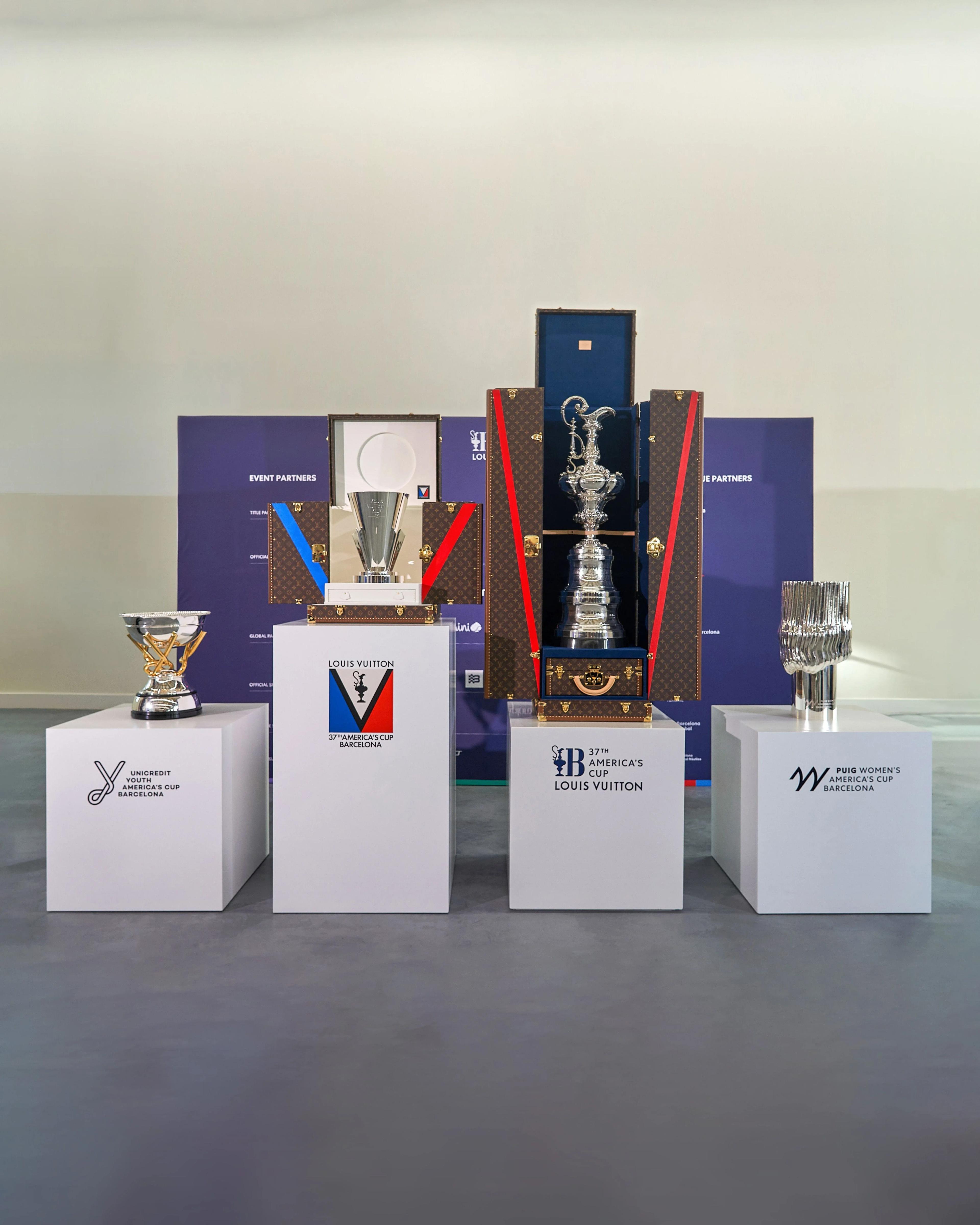 The four trophies of the 37th America’s Cup displayed on white plinths, with branded boxes and trophy cases arranged behind them.